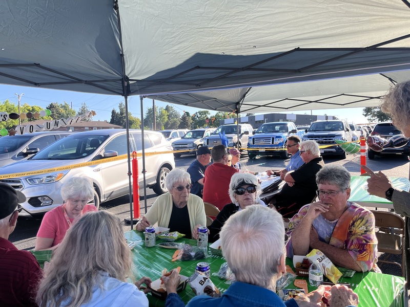 Several older adults are seated at a table under a tent at an outdoor tailgate party.