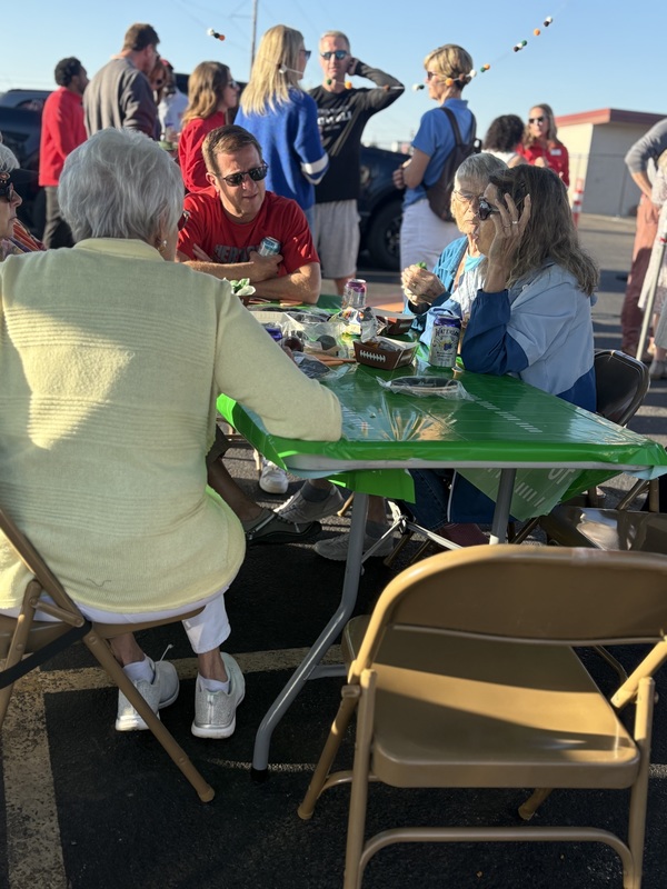 Adults, including Superintendent Todd Lambert, socialize and eat at a table under a tent at an outdoor tailgate party.
