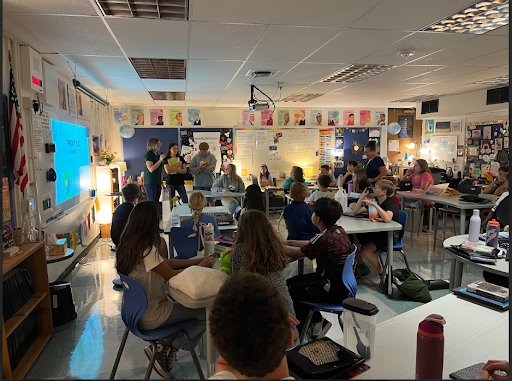 Students are seated at tables in a brightly decorated classroom while a small group and a teacher stand near a projector screen in the front for a presentation or lesson.