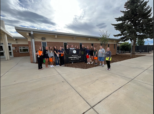 A group of about 15 students and adults holding clipboards or papers stand outside a brick school building under a cloudy sky, posing with a banner for Goddard Middle School.