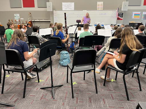 A music class with a teacher standing in front of a whiteboard with sheet music lines, leading a group of middle school students seated on black chairs with music stands.