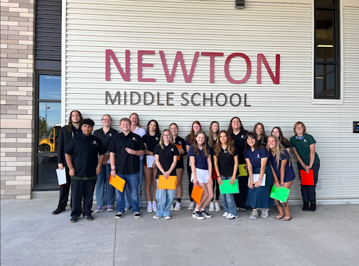 A group of students and a teacher stand smiling outside a building with the large red letters that read NEWTON MIDDLE SCHOOL on a light-colored corrugated wall.