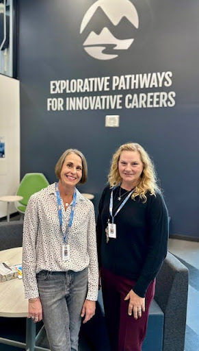  Two women, one with short dark hair and one with long blonde hair, stand in an office lobby with a wall sign that reads "EXPLORATIVE PATHWAYS FOR INNOVATIVE CAREERS."
