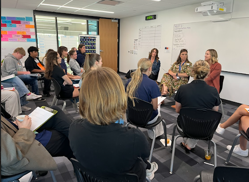  A group of students are seated in a classroom, listening to a panel of three women who are seated in front of a whiteboard during a presentation or discussion.