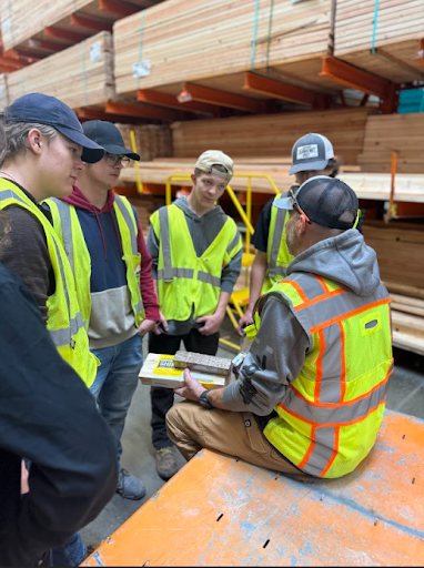  Five young men in high-visibility vests are listening to a man wearing a vest and safety glasses while seated on a pallet jack, discussing building materials in a warehouse or home improvement store.