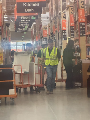 Two adults and two students in high-visibility vests are moving a cart loaded with a white object down an aisle of a home improvement store.
