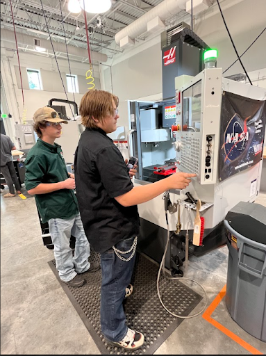 Two male students stand next to a HAAS CNC machine with a NASA banner, one operating the control panel and the other observing.