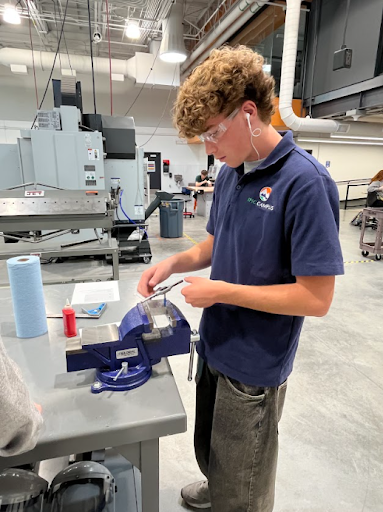 A male student wearing safety glasses and an EPIC Campus polo shirt is working with a small tool and a bench vise on a workbench in a bright machine shop.