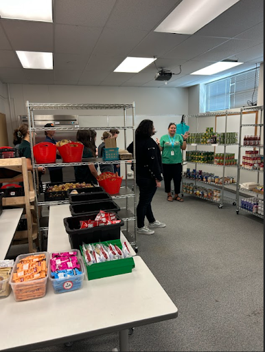 A group of people are gathered in a room that looks like a food pantry, with tables displaying snacks and shelves stocked with canned goods.