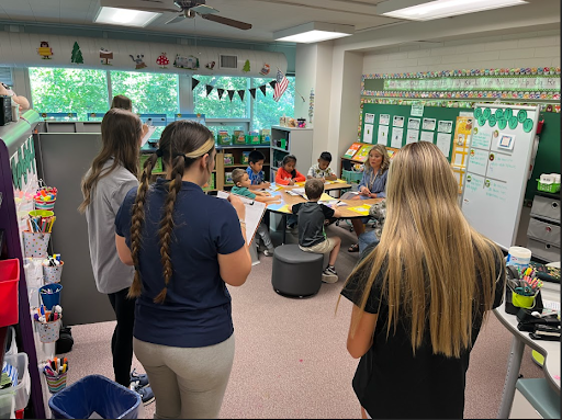 Three adult women observe a teacher and a small group of elementary school students seated at a round table working on a lesson in a colorful classroom.