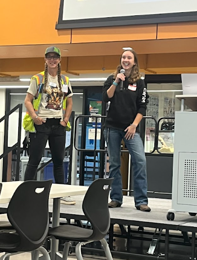 Two women are standing on a stage; one in a high-vis vest and cap, the other in a black hoodie and jeans smiling while holding a microphone.