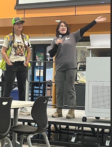 Two women are standing on a stage; one in a high-vis vest and cap, the other in a gray shirt speaking into a microphone and gesturing with her arm.