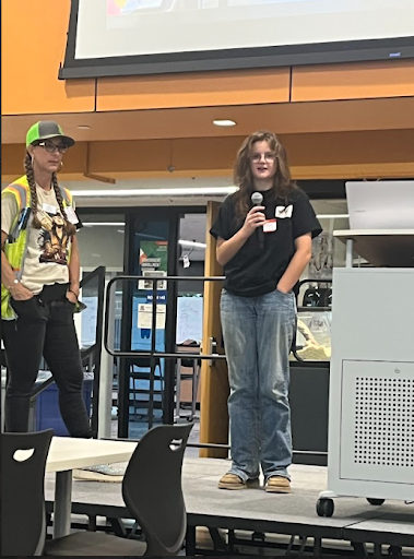 Two women are standing on a stage; one in a high-vis vest, the other in a black shirt and jeans speaking into a microphone.