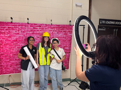 Three students in construction gear (hard hats, vests, safety glasses) pose for a photo in front of a bright pink brick backdrop. A fourth person takes the photo using a phone and a large ring light.