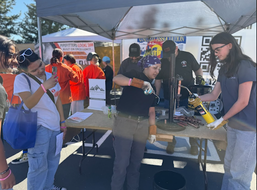 Students at an outdoor event watch a demonstration under a tent. Two women are demonstrating pipe soldering with a torch, while another student takes a picture.