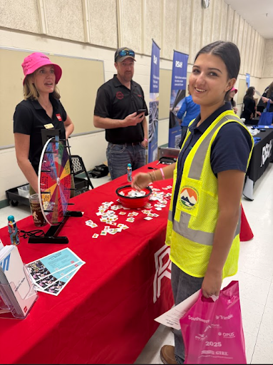  A female student wearing a high-visibility vest and carrying a pink bag stands at a recruitment fair booth. Two adults stand behind a table with a prize wheel and a red tablecloth.