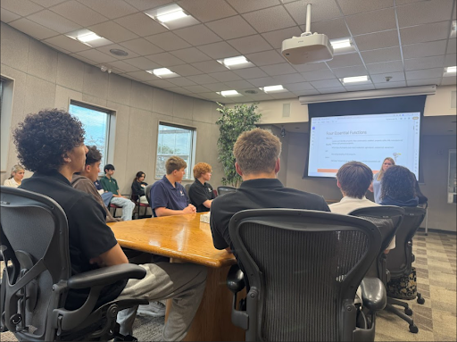  A group of high school students is seated around a conference table, looking at a projector screen that displays a presentation on "Four Essential Functions." A teacher is standing near the screen.