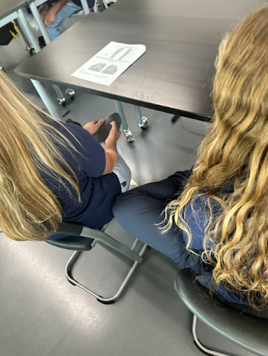 An overhead view of two people with long blonde hair seated at a table. One person is holding a gray sock or foam piece, possibly for a practical lesson. A worksheet is on the table.