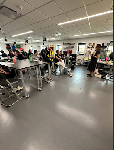 A wide shot of a classroom shows a female teacher in a black dress speaking to a group of students seated at black tables. Exercise equipment is visible along the back wall.