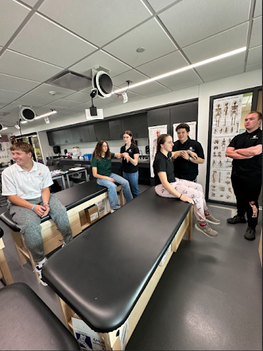 A small group of five students is seated on black treatment tables in a sports medicine lab. Two other students stand to the side as they participate in a practical lesson.