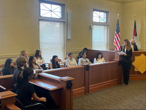  A group of high school students is seated in a courtroom, participating in a mock trial or lesson. A young woman in a black suit stands in the foreground, addressing the students and a man seated in the judge's area.