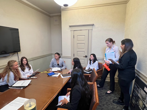  A group of eight students is seated around a polished wooden conference table, discussing documents. Two young women stand at the end of the table, leading the conversation in the meeting room.