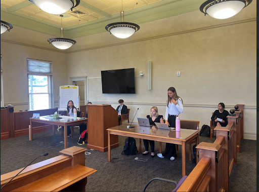  High school students are participating in a mock trial or law class inside a courtroom. Students are seated at wooden tables, with two young women standing and a television screen mounted on the wall.
