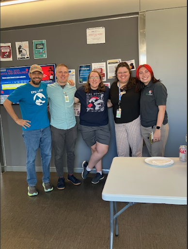  Five young adults—three women and two men—are standing with their arms around each other, smiling at the camera in what looks like an office or break room with informational posters on the wall.