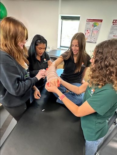 Four students practice applying a wrap and splint to an arm on a training table.