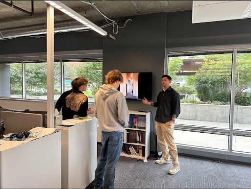 Three people, two students and one instructor, stand in a modern office space discussing a presentation on a screen.