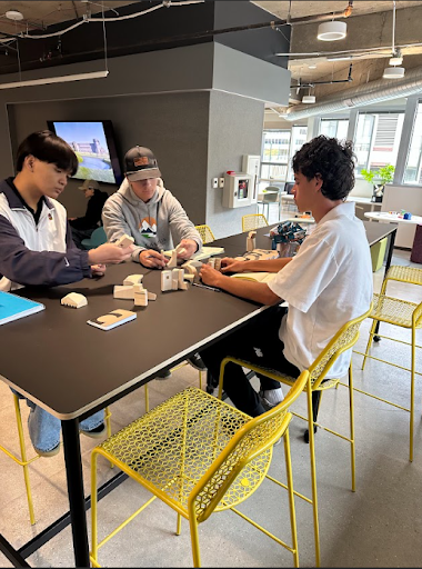 Three students sit at a long table in a modern space, working together to build a structure with small wooden blocks.