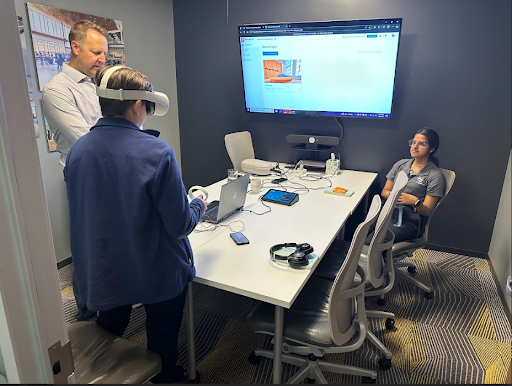 A student wears a VR headset at a table with an instructor and another student, looking at a screen displaying a 3D architectural model.