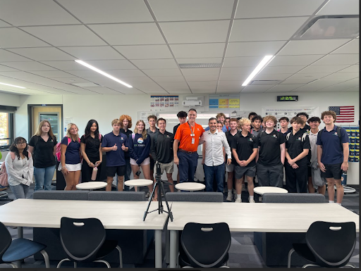 A large group of high school students and two adult men pose for a group photo in a modern classroom with long tables.