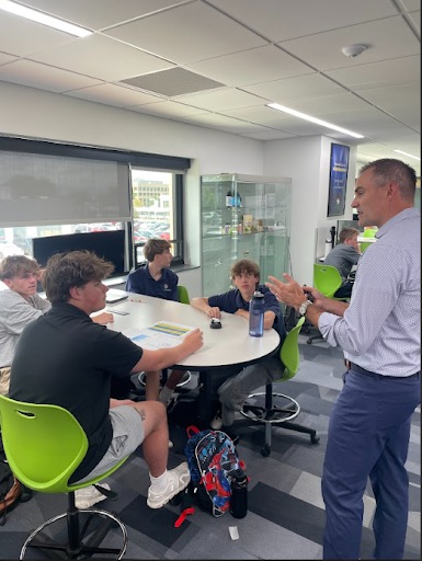 An adult man stands, gesturing while speaking to a small group of four male students seated around a white oval table in a bright, modern classroom.