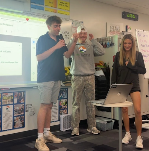 Three young people presenting in a classroom, with one holding a microphone and a laptop on a stand.