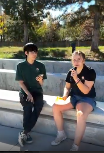Two students sitting outdoors on concrete steps, one holding a microphone as if conducting an interview.