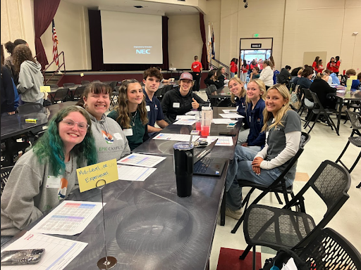 A group of eight high school students sit at a long table in a large room, smiling at the camera during a group event.