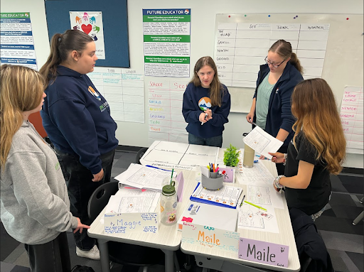 Five female students collaborate around a table covered with papers and school supplies in a classroom.
