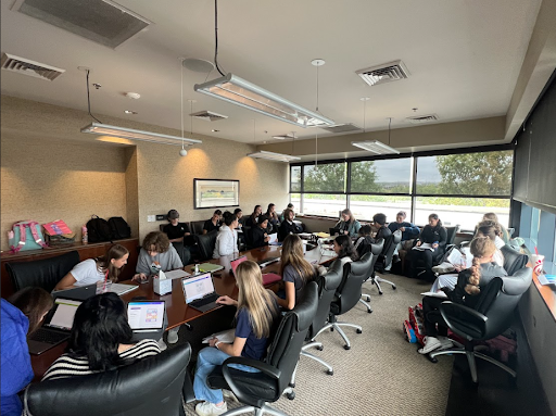 A large group of students are seated around a conference room table for a meeting, with windows along one side.