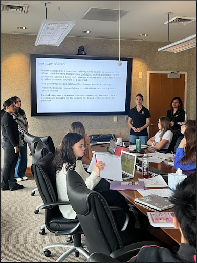 Students presenting a "Summary of event" slide on a large screen to a group seated around a conference table.
