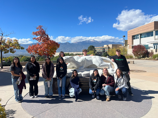 A group of students is posing around a white panther statue outdoors on a sunny day with a snow-capped mountain in the background.