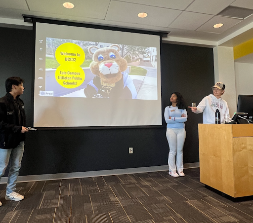 Two students present in front of a large screen displaying a welcome message and a university mascot in a modern lecture room.