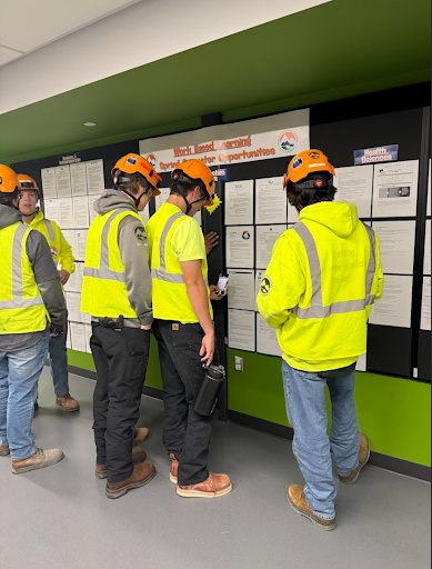 A group of students in safety vests and hard hats stand in front of a wall of "Work-Based Learning" opportunity postings.