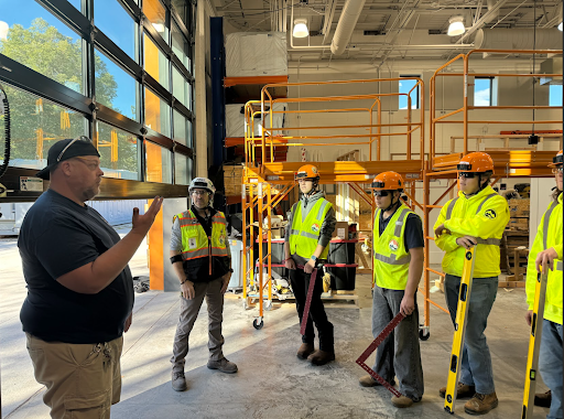 An instructor talks to a group of students wearing safety vests and hard hats in a large indoor construction lab space with scaffolding.