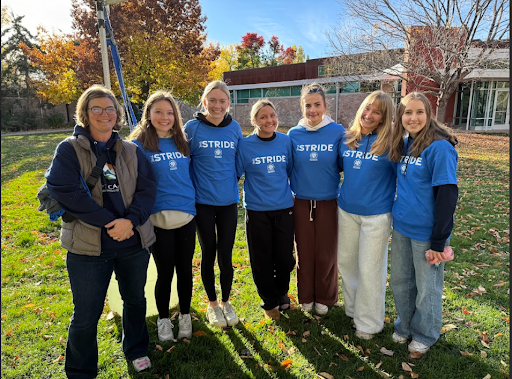 A group of seven women, most wearing matching blue "STRIDE" shirts, stand together outdoors on a grassy lawn with trees and a modern building behind them.