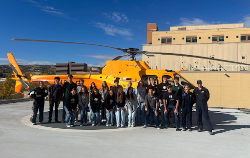 A group of about 20 students and several Flight For Life crew members pose for a photo outdoors in front of an orange helicopter on a sunny day.