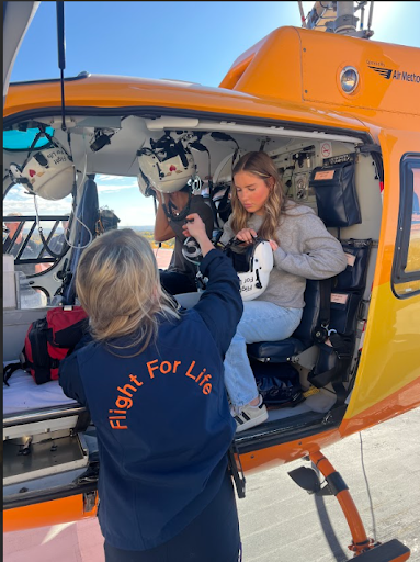 A person in a "Flight for Life" jacket assists a student with putting on a white helmet while seated inside the open door of an orange medical helicopter.