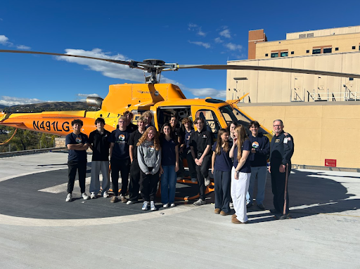 A large group of students and two Flight For Life crew members stand together in front of an orange medical helicopter on a rooftop landing pad.