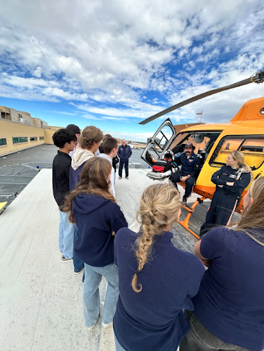 tudents on a rooftop helipad listen to a Flight For Life crew member sitting in the open door of a bright orange helicopter, with a wide view of the sky and city buildings.