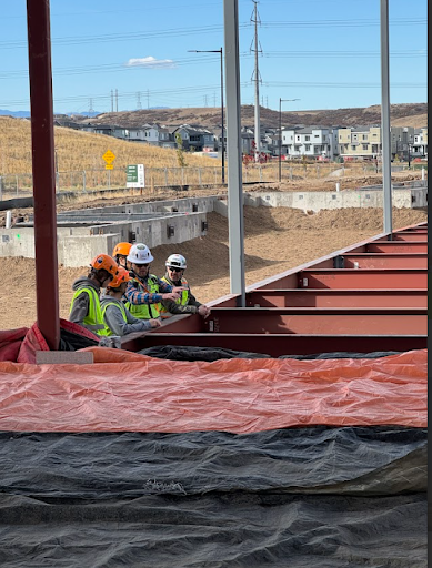 Four workers in hard hats and safety vests are gathered and talking at the edge of a construction site, with a large steel structure and suburban housing visible behind them.